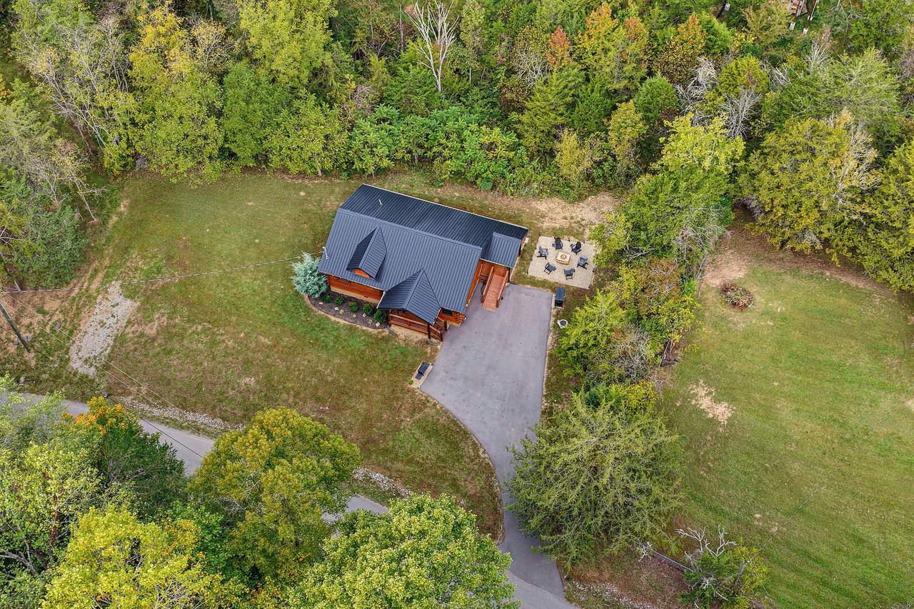 An aerial view showcases the property nestled in a peaceful natural setting surrounded by lush trees and open lawn space. A private patio area and ample parking complete this charming wooded retreat.