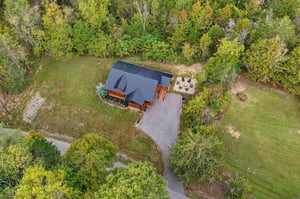 An aerial view showcases the property nestled in a peaceful natural setting surrounded by lush trees and open lawn space. A private patio area and ample parking complete this charming wooded retreat.