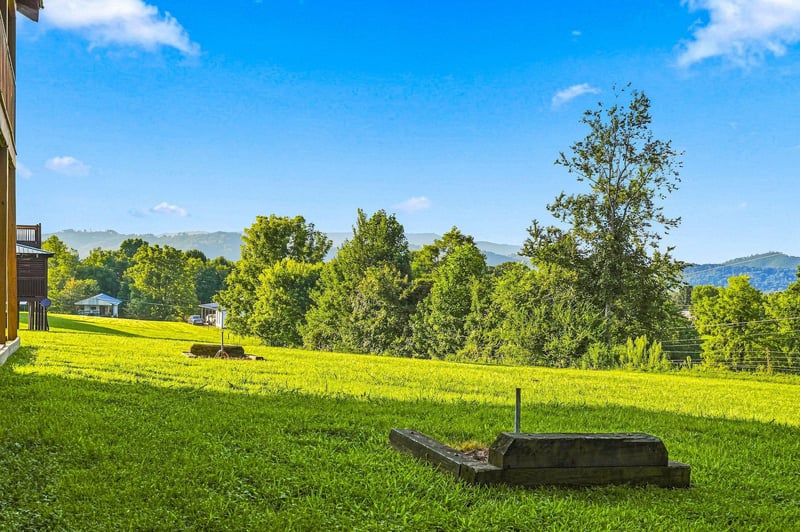 Spacious outdoor grounds with rolling green lawn, surrounded by mature trees and distant mountain views under a bright blue sky.