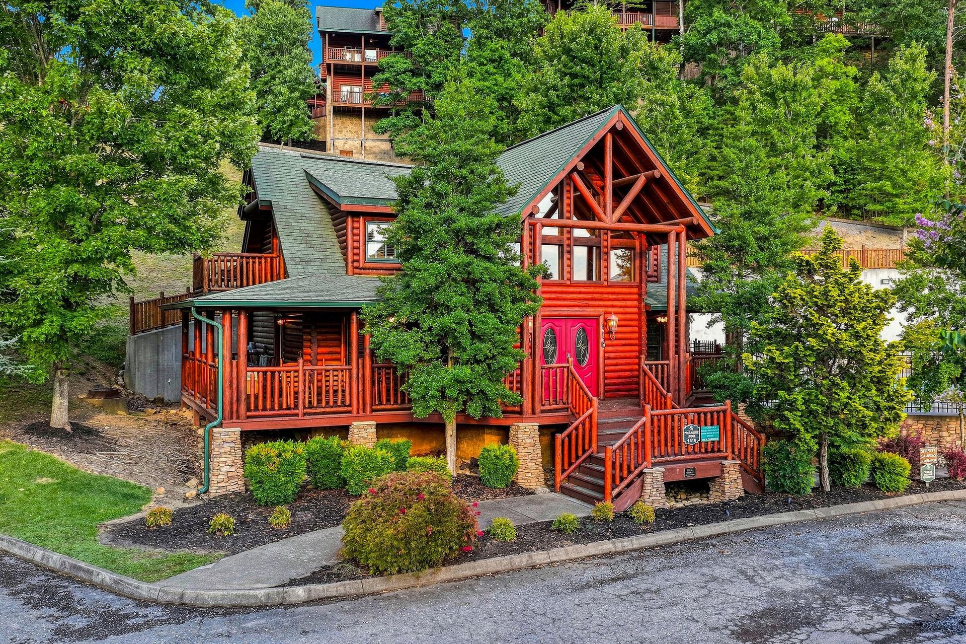Charming log cabin nestled in lush wooded surroundings offers a peaceful mountain retreat with distinctive red accents, multiple decks, and inviting architecture surrounded by vibrant landscaping.