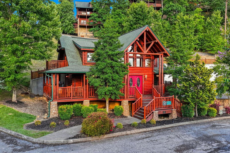 Charming log cabin nestled in lush wooded surroundings offers a peaceful mountain retreat with distinctive red accents, multiple decks, and inviting architecture surrounded by vibrant landscaping.