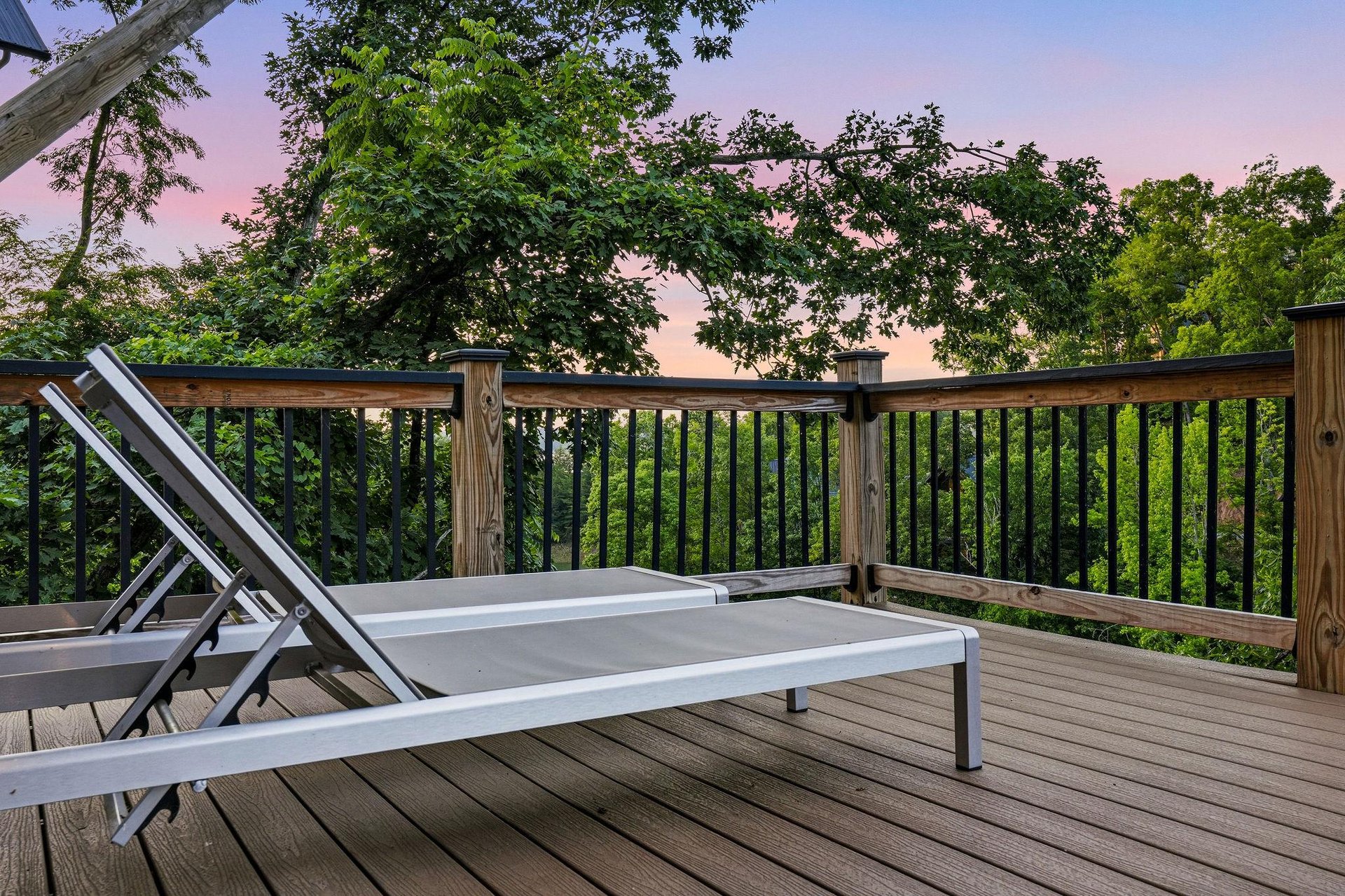 A peaceful deck overlooks lush treetops as the sky glows with soft pink and purple hues at sunset, offering your perfect spot to unwind with nature.