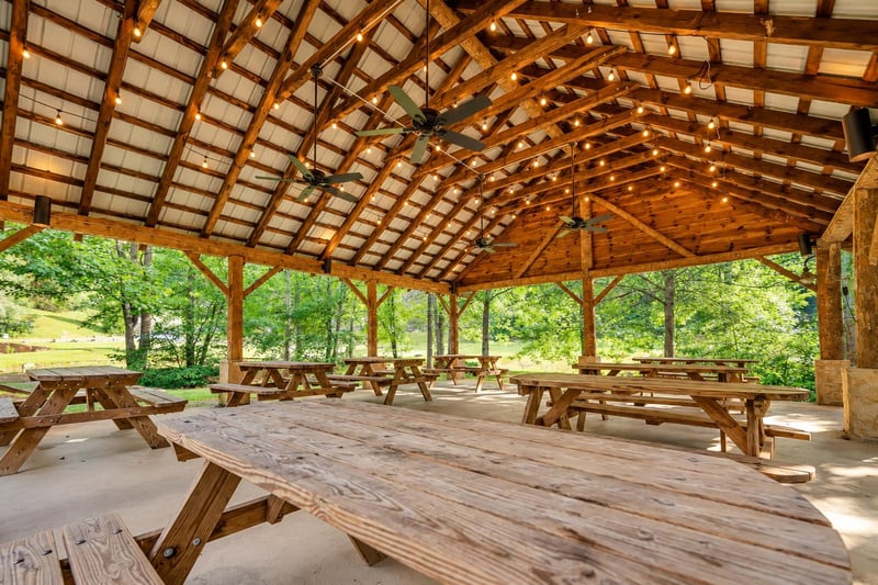 A spacious openair pavilion surrounded by lush greenery, featuring rustic picnic tables beneath a beautifully crafted vaulted wood ceiling strung with string lights—ideal for group gatherings and out
