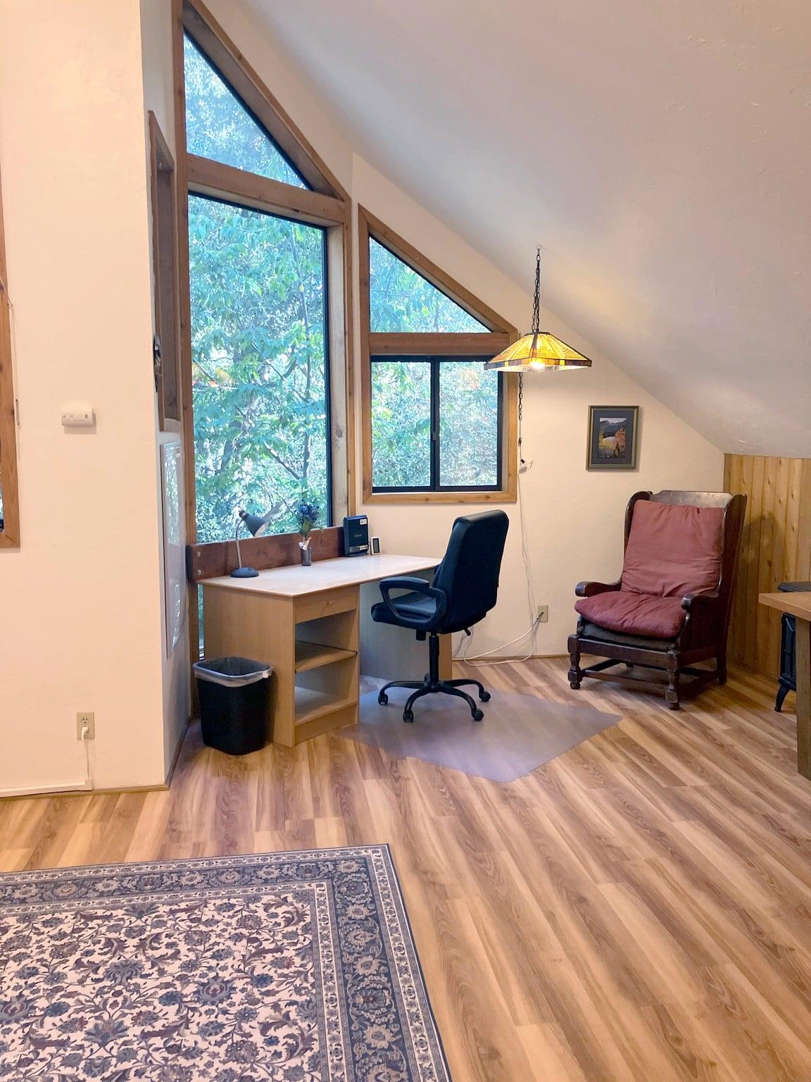 A dedicated workspace tucked into the primary bedroom, with a desk, ergonomic chair, and big windows looking out into the trees — perfect for anyone who needs to get a little work done during their stay.