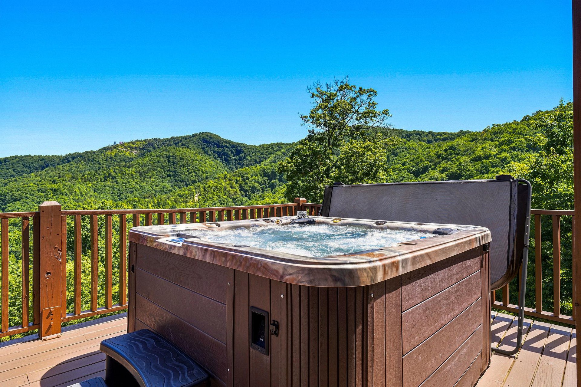 A hot tub on your private deck overlooks lush, rolling mountains under brilliant blue skies—perfect for unwinding in the bubbling water while soaking in stunning natural views.