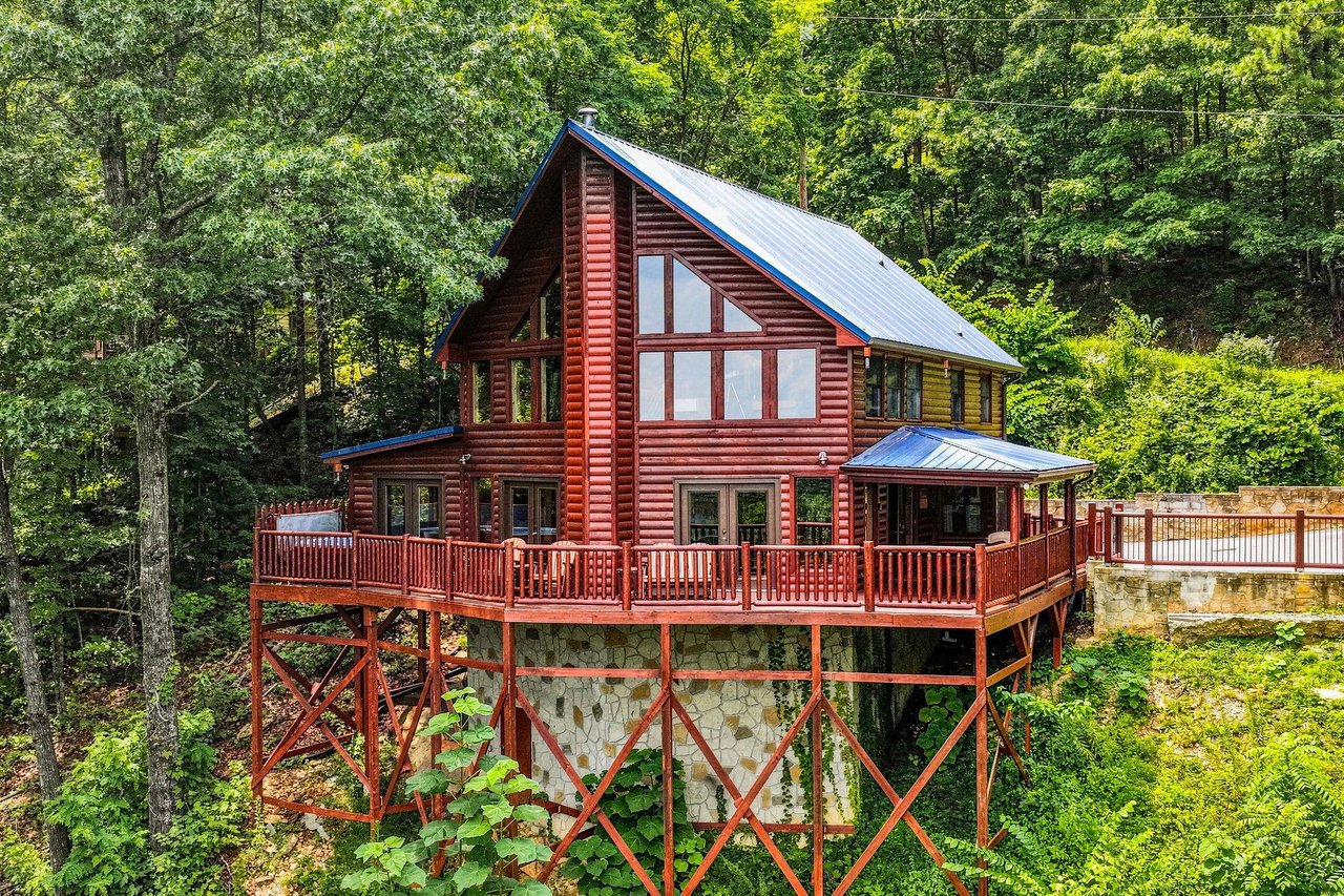 A welcoming log cabin nestled among lush forest trees, featuring a spacious wraparound deck and large windows that invite natural light into your mountain retreat.