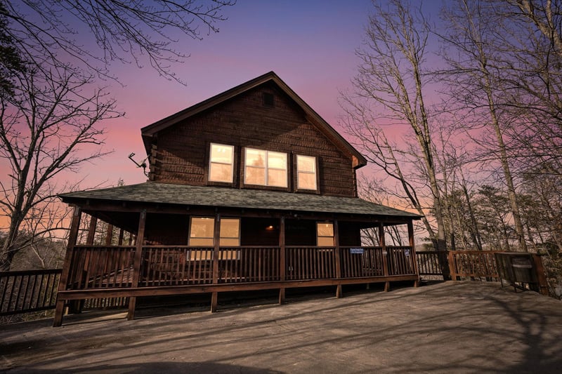 Charming cabin with wraparound deck nestled among trees, featuring warm sunset lighting and peaceful forest surroundings.