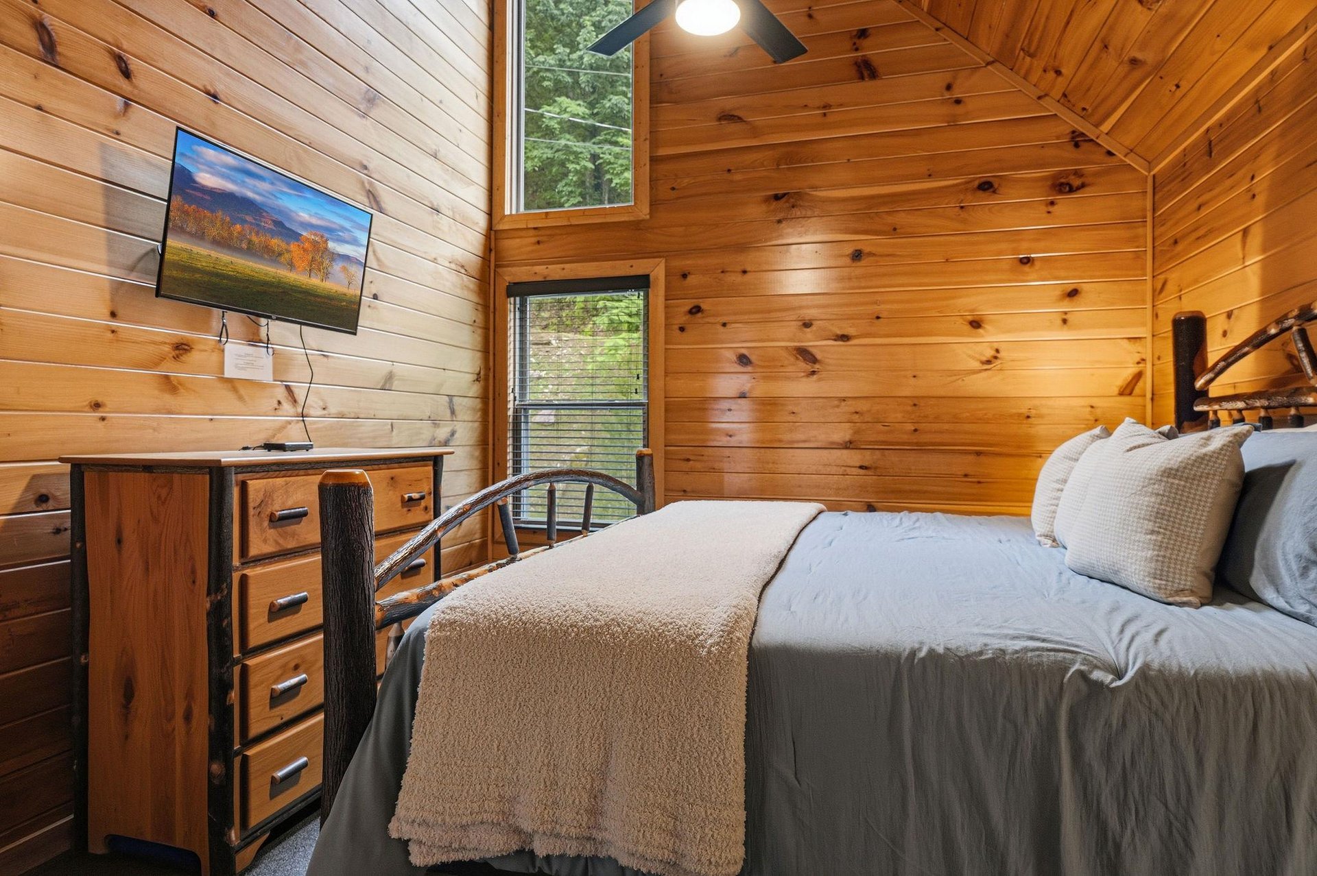 Drift off in your cozy cabin bedroom, where warm wood surrounds you, high ceilings open the space, and treetop views peek through the windows—your peaceful retreat awaits.