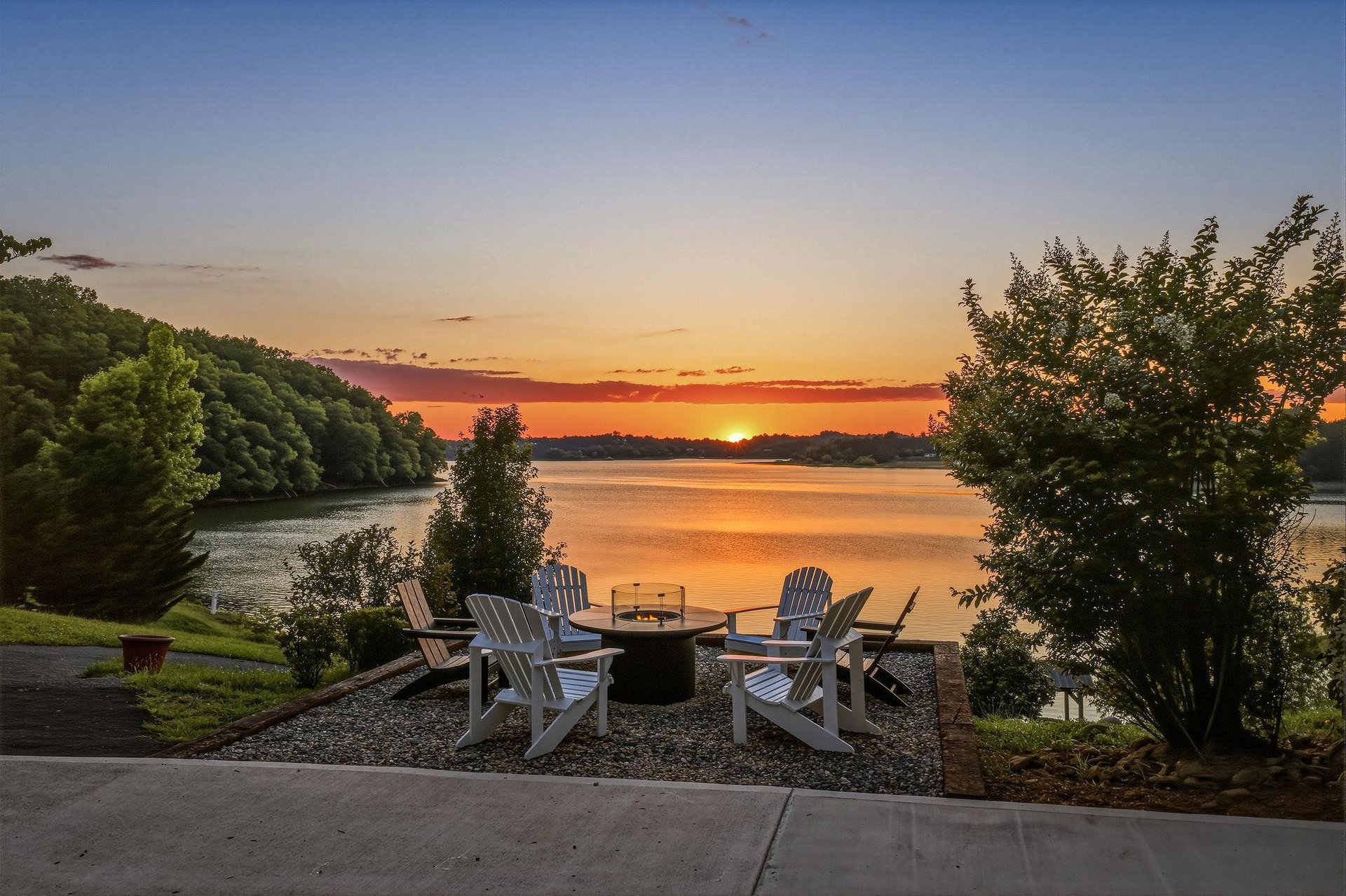 A peaceful lakefront setting with Adirondack chairs and fire pit, framed by lush trees as the sun sets over calm waters.