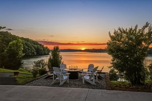 A peaceful lakefront setting with Adirondack chairs and fire pit, framed by lush trees as the sun sets over calm waters.