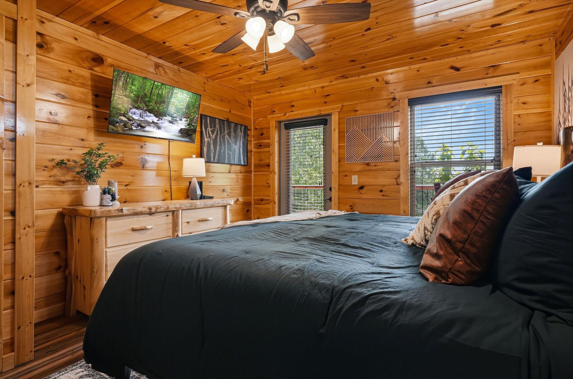 Sink into the plush bed beneath warm pine ceilings and ceiling fan. Natural light fills your cozy retreat, with forest views framing a peaceful nights rest.