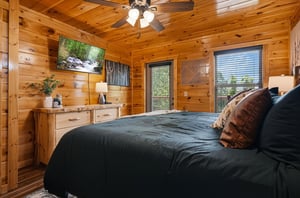 Sink into the plush bed beneath warm pine ceilings and ceiling fan. Natural light fills your cozy retreat, with forest views framing a peaceful nights rest.