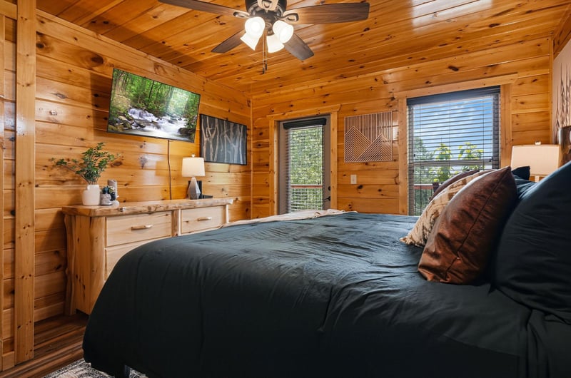 Sink into the plush bed beneath warm pine ceilings and ceiling fan. Natural light fills your cozy retreat, with forest views framing a peaceful nights rest.