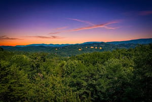 Stunning sunset over forested mountains and distant lit cabins, captured from the propertys scenic surroundings—an idyllic natural backdrop for a mountain getaway.