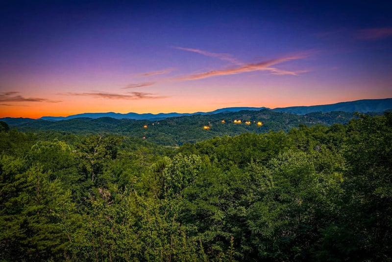 Stunning sunset over forested mountains and distant lit cabins, captured from the propertys scenic surroundings—an idyllic natural backdrop for a mountain getaway.
