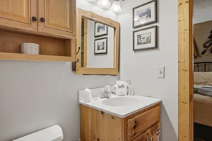 Your private bathroom features warm wood cabinetry, a clean vanity stocked with fresh amenities, and charming mountaininspired artwork on the walls, creating a cozy alpine retreat feel.