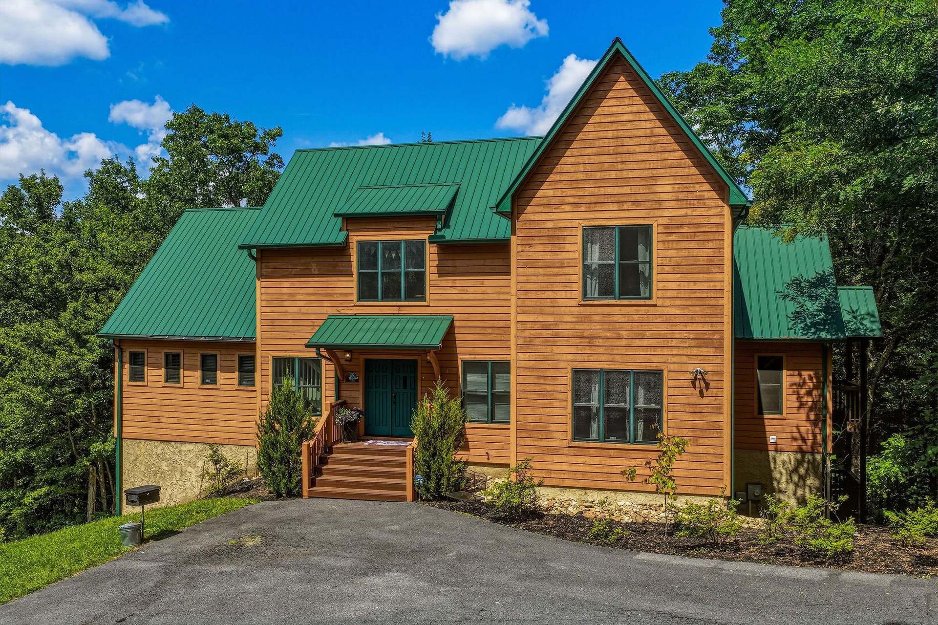 A charming woodsided retreat with a distinctive green metal roof, nestled among tall trees in a peaceful, natural setting with convenient paved parking.