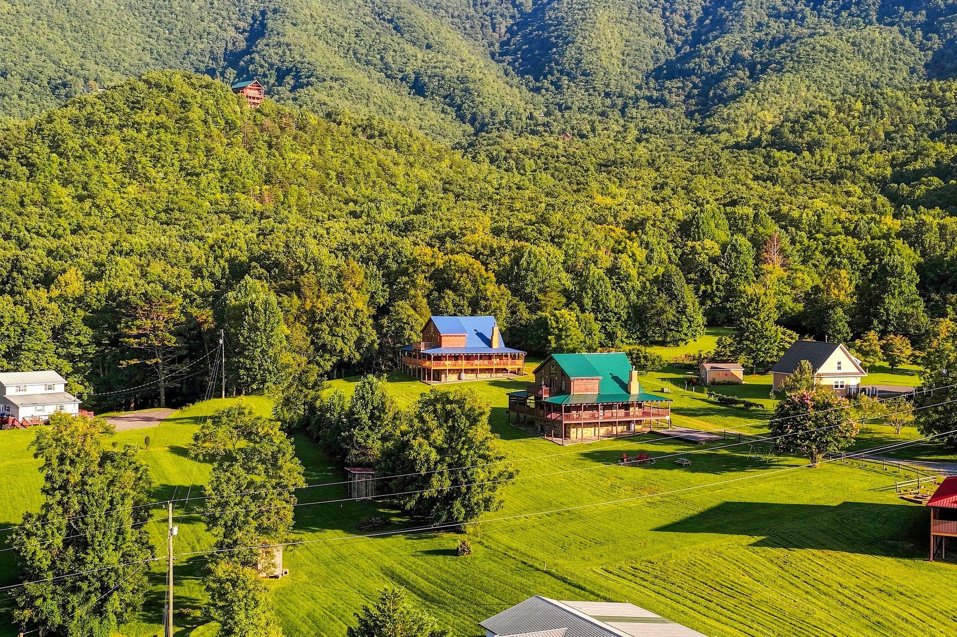 An aerial view captures the rental property nestled among lush forestcovered mountains, surrounded by vibrant green lawns and mature trees in a peaceful countryside setting.