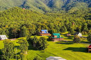 An aerial view captures the rental property nestled among lush forestcovered mountains, surrounded by vibrant green lawns and mature trees in a peaceful countryside setting.