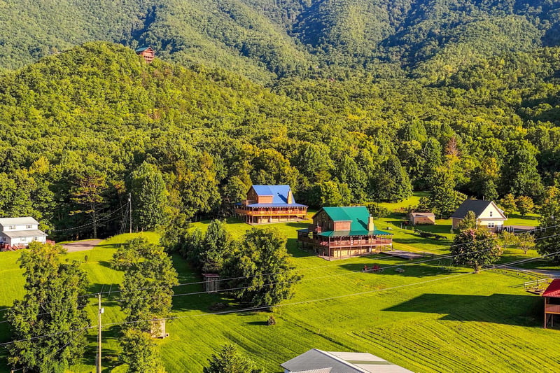 An aerial view captures the rental property nestled among lush forestcovered mountains, surrounded by vibrant green lawns and mature trees in a peaceful countryside setting.