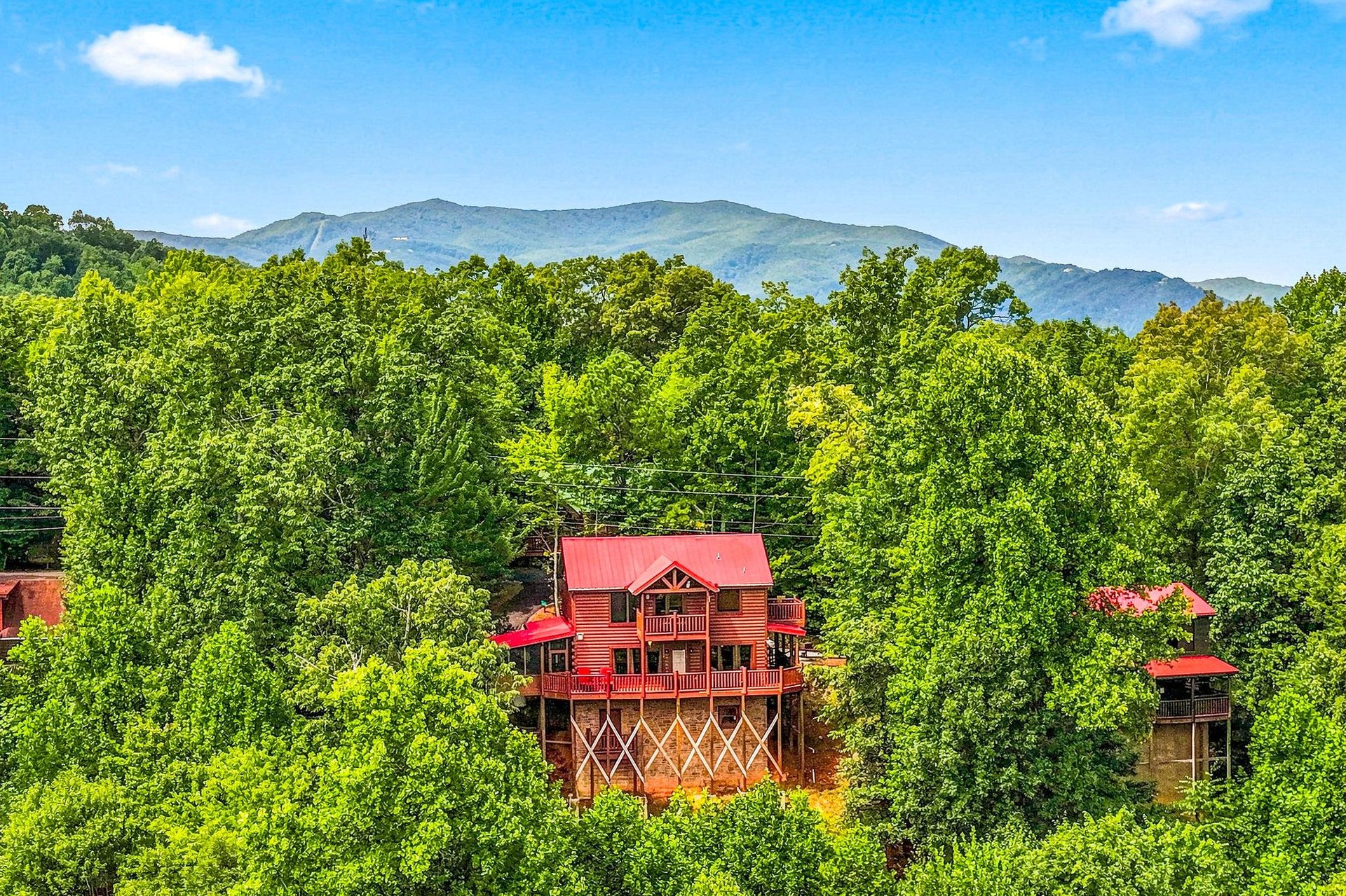 A charming mountain cabin with a distinctive red roof nestled among lush forest, with stunning mountain ridges rising majestically in the distance under a clear blue sky.