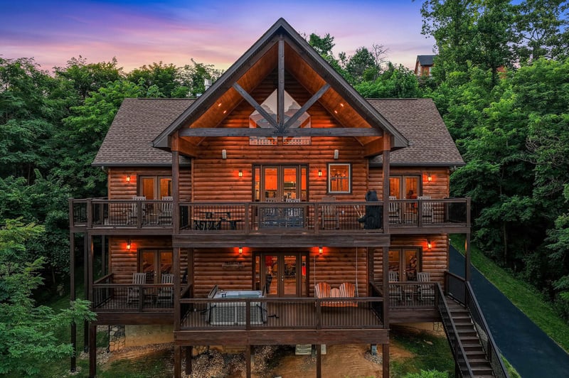 A striking log cabin exterior stands illuminated at dusk, nestled among lush trees and hillside greenery, showcasing rustic architecture and spacious multilevel decks perfect for mountain getaways.