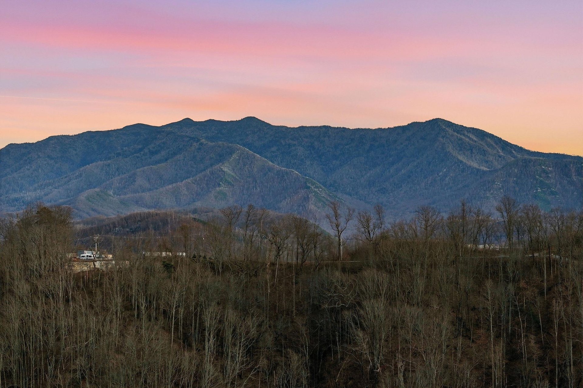 Stunning mountain landscape with vibrant sunset colors painting the sky in soft pinks and purples above rolling forested hills.