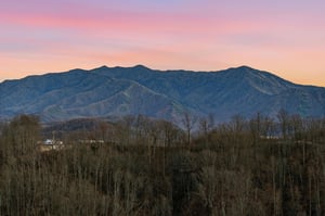 Stunning mountain landscape with vibrant sunset colors painting the sky in soft pinks and purples above rolling forested hills.