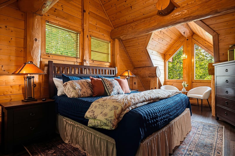 Drift off in this cozy cabin bedroom, where warm wood beams, soft bedside lighting, and treetop views through charming windows invite peaceful sleep.