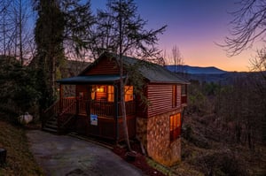 A charming log cabin nestled in the mountains, perfectly positioned to showcase stunning sunset views over the surrounding peaks and valleys.