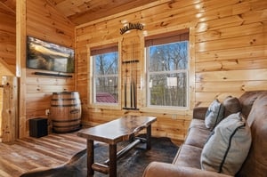 Relax in your rustic living room with warm wood walls, cozy seating, and peaceful forest views through large windows.