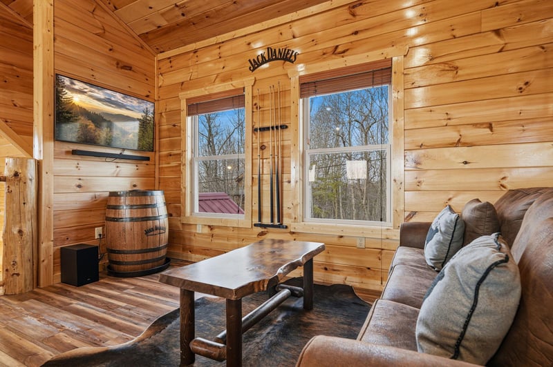 Relax in your rustic living room with warm wood walls, cozy seating, and peaceful forest views through large windows.