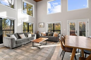 Step into this stunning living area, where soaring ceilings and floortoceiling windows frame breathtaking autumn foliage. Sink into plush gray sofas, gather around the dining table, and feel the war