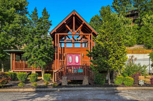 Charming log cabin nestled among evergreens with distinctive pink double doors and an open upper balcony. The Aframe design and natural timber construction provide a welcoming mountain retreat in a p