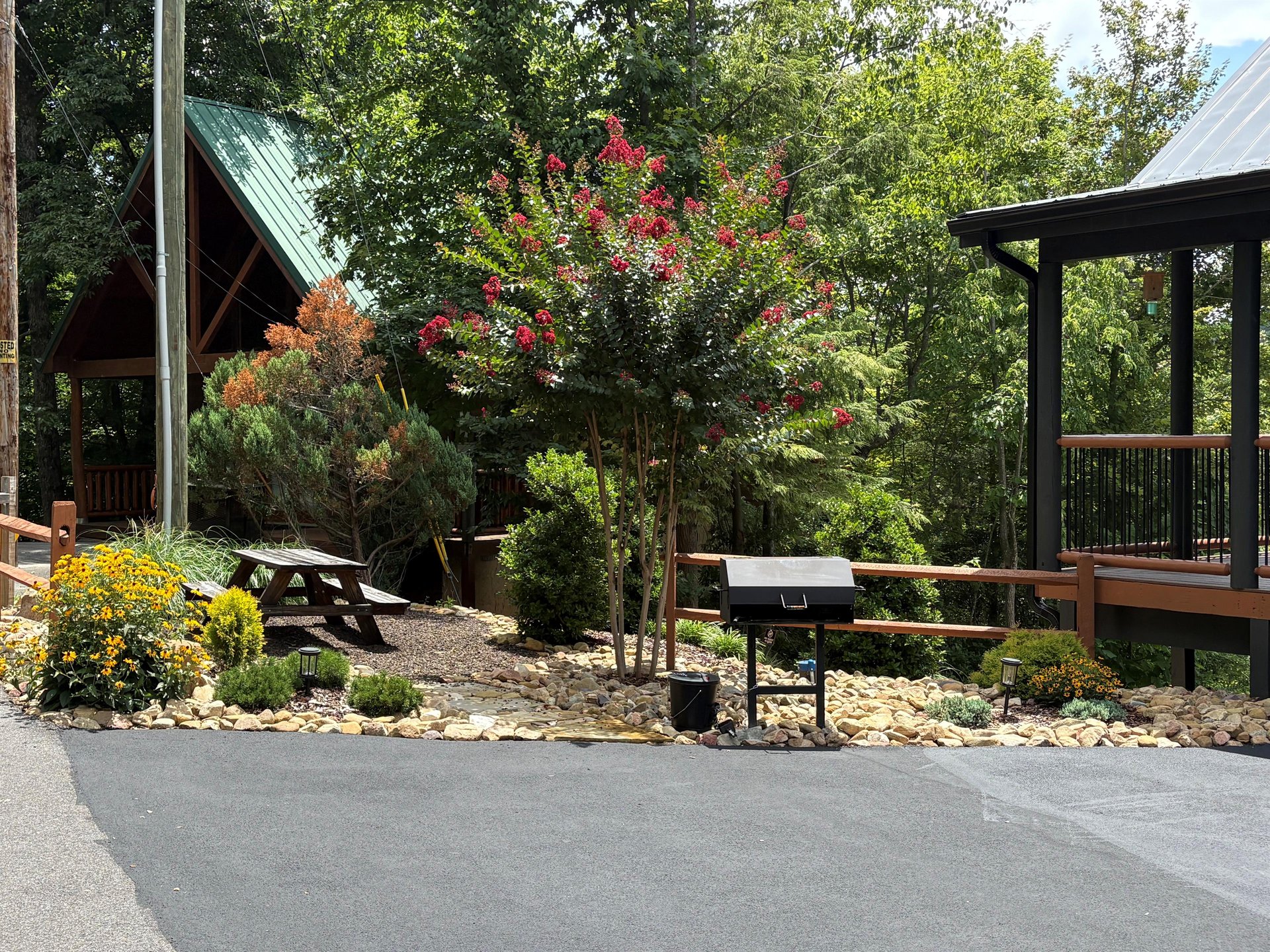 Nestled in lush landscaping with blooming trees and colorful flowers, the property features two charming cabin structures with paved parking, a picnic table, and a BBQ grill for outdoor gatherings.