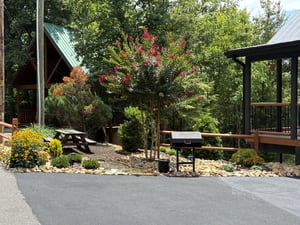 Nestled in lush landscaping with blooming trees and colorful flowers, the property features two charming cabin structures with paved parking, a picnic table, and a BBQ grill for outdoor gatherings.