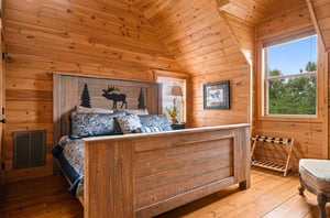 Step into your cozy cabin retreat with vaulted wood ceilings and rustic charm. Your bed features a handcrafted headboard, and natural light pours through the windows to frame peaceful treetop views.