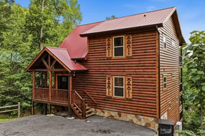 A charming log cabin nestled in lush greenery, featuring a welcoming red metal roof and covered porch. The warm wood tones and mountain setting create a peaceful retreat.