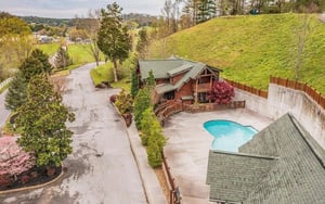 Stunning aerial view of the rustic cabin retreat nestled in rolling green hills, featuring a private swimming pool and surrounded by lush spring landscapes.