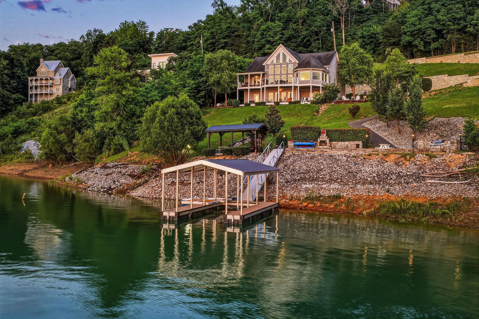 Stunning lakefront property features private boat dock, terraced landscaping, and covered outdoor entertaining area surrounded by lush wooded hillside in peaceful waterfront community.