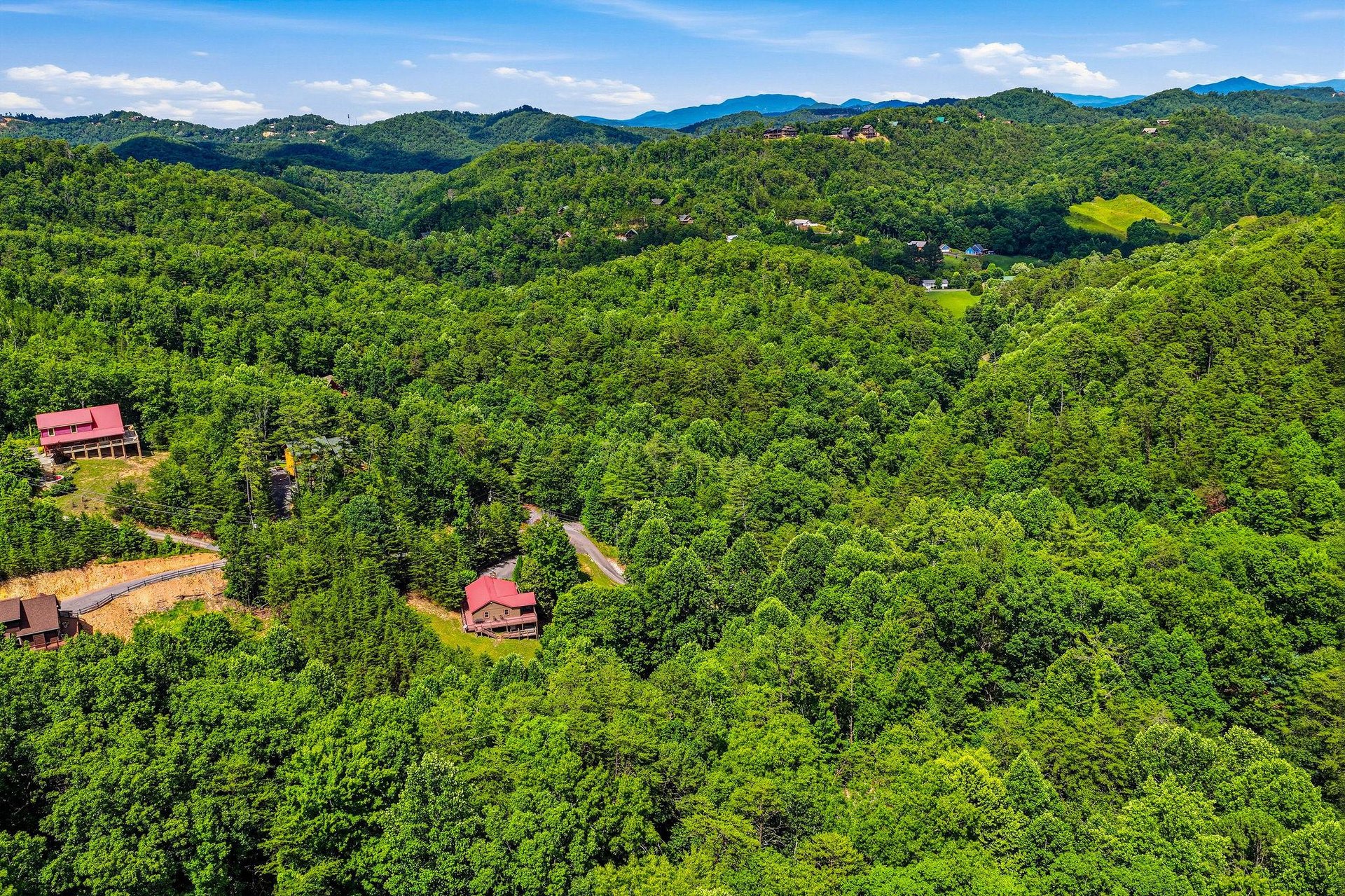 Aerial view of the property nestled in rolling forested mountains, featuring redroofed buildings along winding roads through lush green valleys and distant peaks.