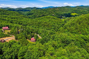 Aerial view of the property nestled in rolling forested mountains, featuring redroofed buildings along winding roads through lush green valleys and distant peaks.