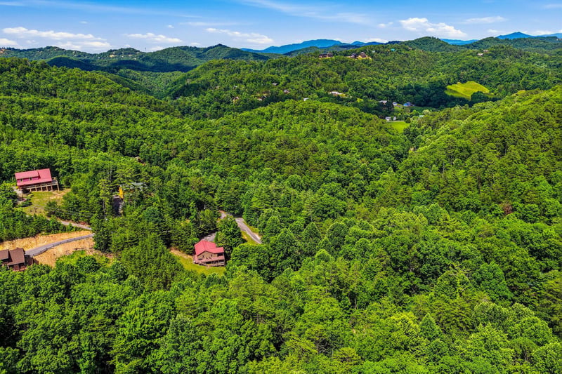 Aerial view of the property nestled in rolling forested mountains, featuring redroofed buildings along winding roads through lush green valleys and distant peaks.