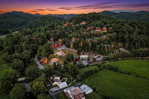 Aerial view captures a charming mountain resort nestled in lush forests, with vibrant sunlit cabins and scenic peaks framing the peaceful landscape at dusk.