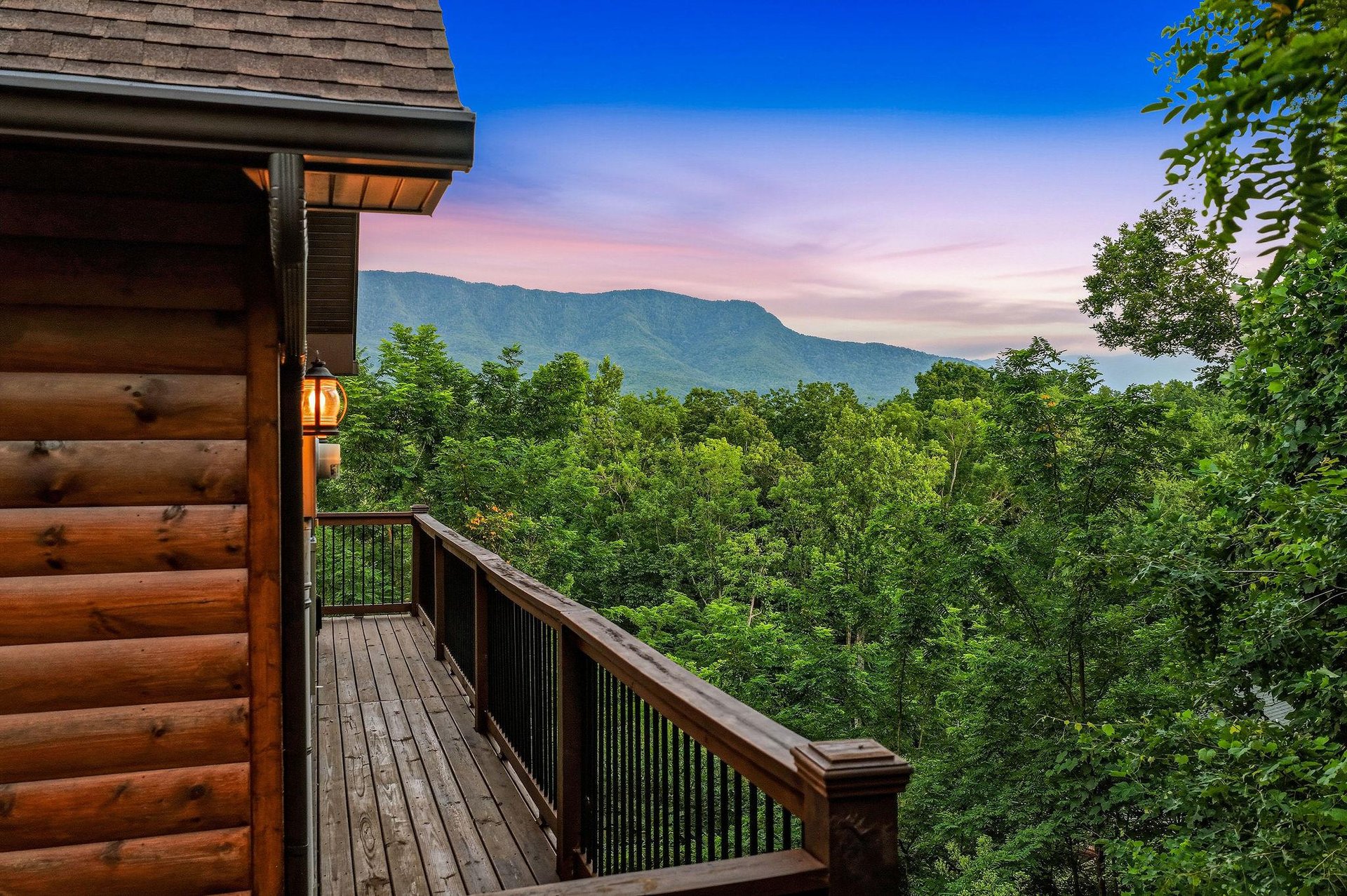 Property nestled in a mountainous area with lush forest surroundings and scenic natural landscape. The deck overlooks rolling green hills and distant peaks under a colorful evening sky.