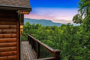 Property nestled in a mountainous area with lush forest surroundings and scenic natural landscape. The deck overlooks rolling green hills and distant peaks under a colorful evening sky.