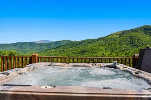 A stunning mountain vista unfolds beyond your private hot tub, where lush green peaks stretch endlessly beneath a clear blue sky—the perfect setting for soaking away stress.