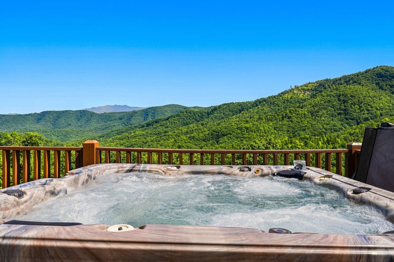 A stunning mountain vista unfolds beyond your private hot tub, where lush green peaks stretch endlessly beneath a clear blue sky—the perfect setting for soaking away stress.