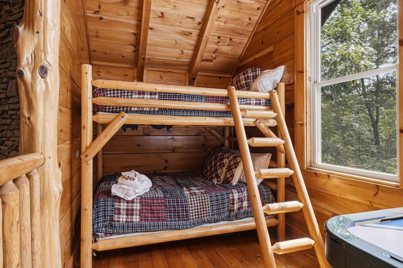 Your cozy cabin bedroom features handcrafted log bunk beds with soft plaid linens, perfect for family bonding. Natural light streams through the window, framing peaceful forest views right from your r
