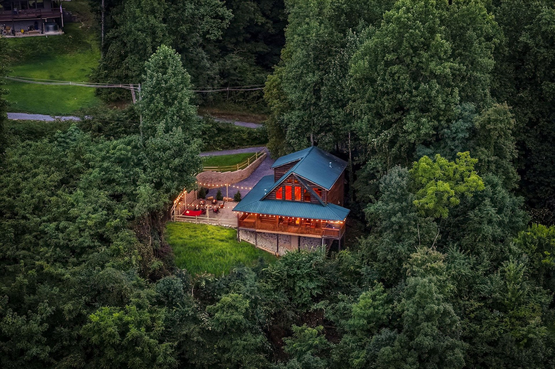 Aerial view showcases a charming log cabin with a distinctive blue metal roof and red accents, nestled privately among lush forest greenery with an inviting deck for outdoor relaxation.